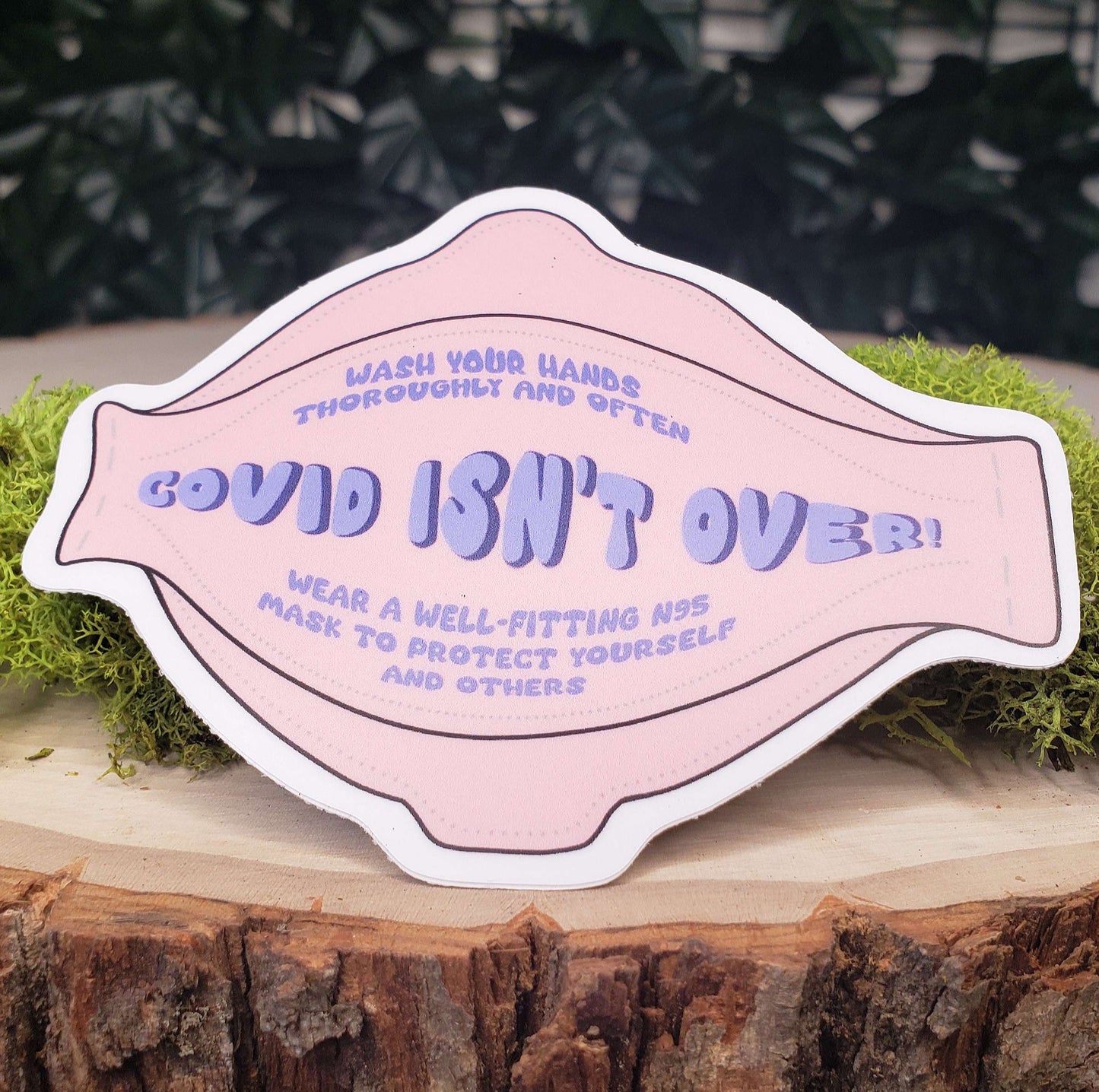 Sticker of a pink n95 respirator mask with large purple bubble letters across the middle reading "Covid Isn't Over." In smaller letters above and below it reads "Wash your hands thoroughly and often" and "wear a well-fitting n95 mask to protect yourself and others. the sticker is sitting on moss and wood with ivy leaves in the background