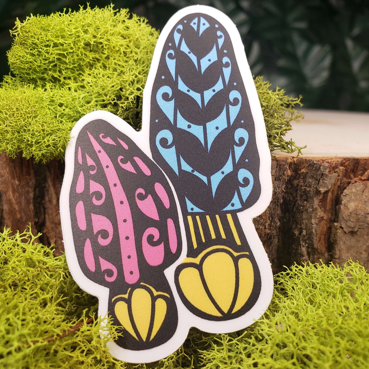 Sticker of pink, blue, and yellow morel mushrooms with stark black line art reminiscent of henna tattoo art. the sticker is sitting on moss and wood with ivy leaves in the background