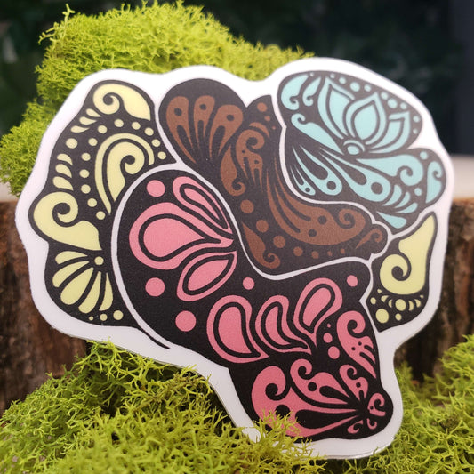 Sticker of brown and pastel yellow, red, turquoise witches' butter mushrooms with stark black line art reminiscent of henna tattoo art. the sticker is sitting on moss and wood with ivy leaves in the background