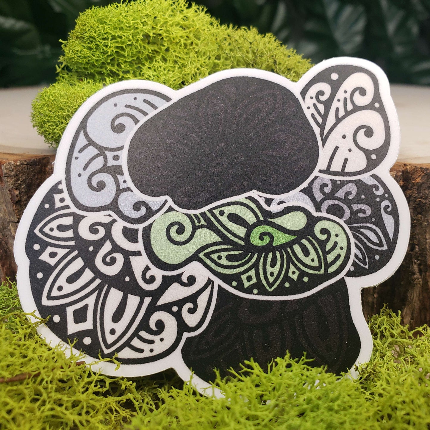 Sticker of green, cream, gray, and black hen of the woods mushrooms with stark black line art reminiscent of henna tattoo art. the sticker is sitting on moss and wood with ivy leaves in the background