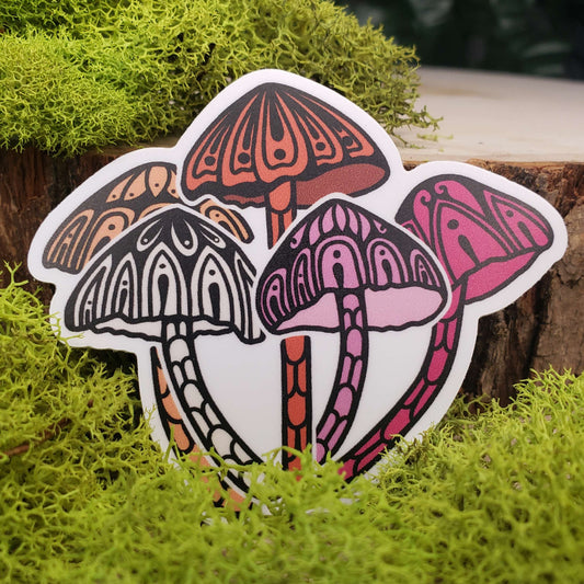 Sticker of light orange, dark orange, light pink, dark pink, and cream liberty cap mushrooms with stark black line art reminiscent of henna tattoo art. the sticker is sitting on moss and wood with ivy leaves in the background