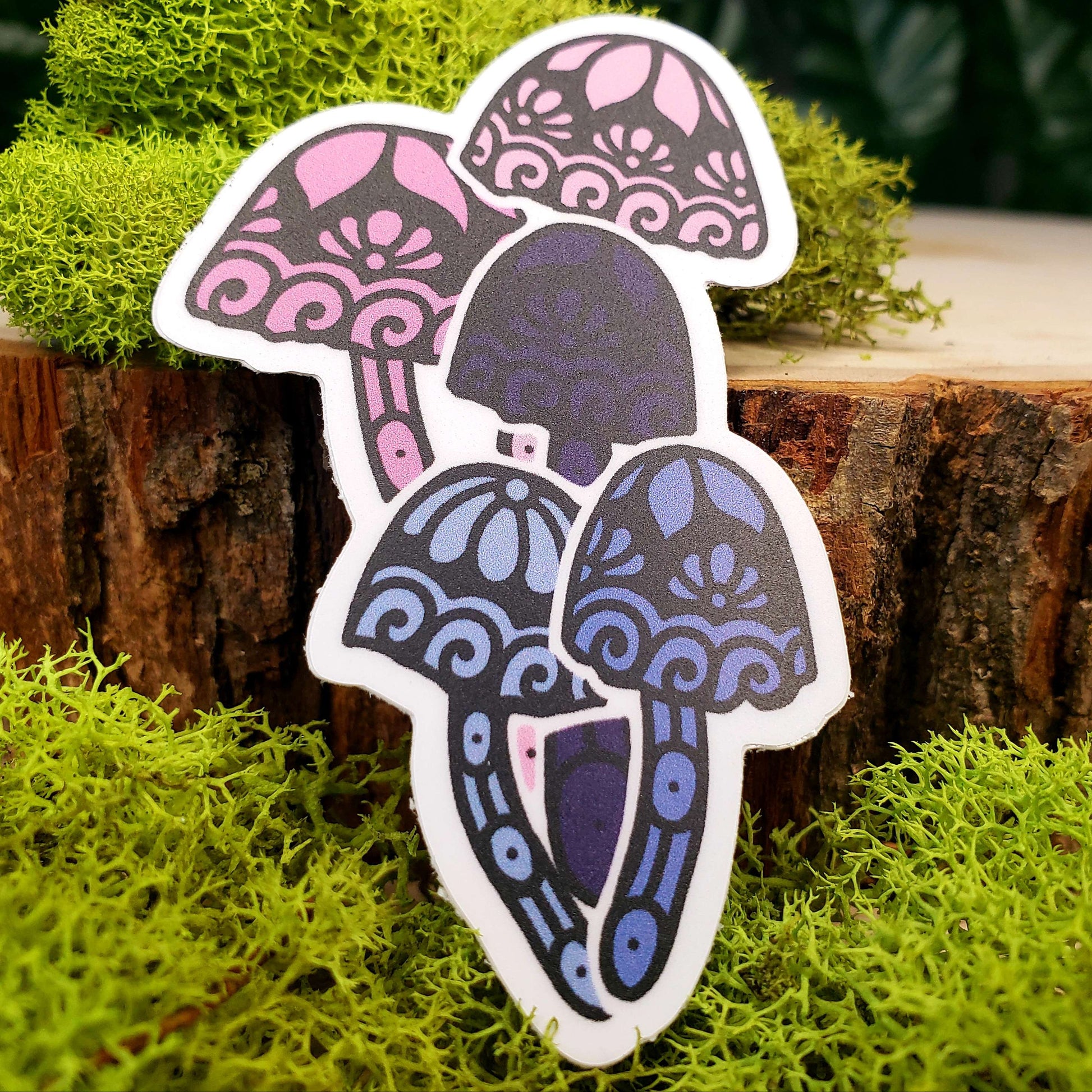 Sticker of pastel pink, medium pink, pastel blue, medium blue, and dark blue mica cap mushrooms with stark black line art reminiscent of henna tattoo art. the sticker is sitting on moss and wood with ivy leaves in the background