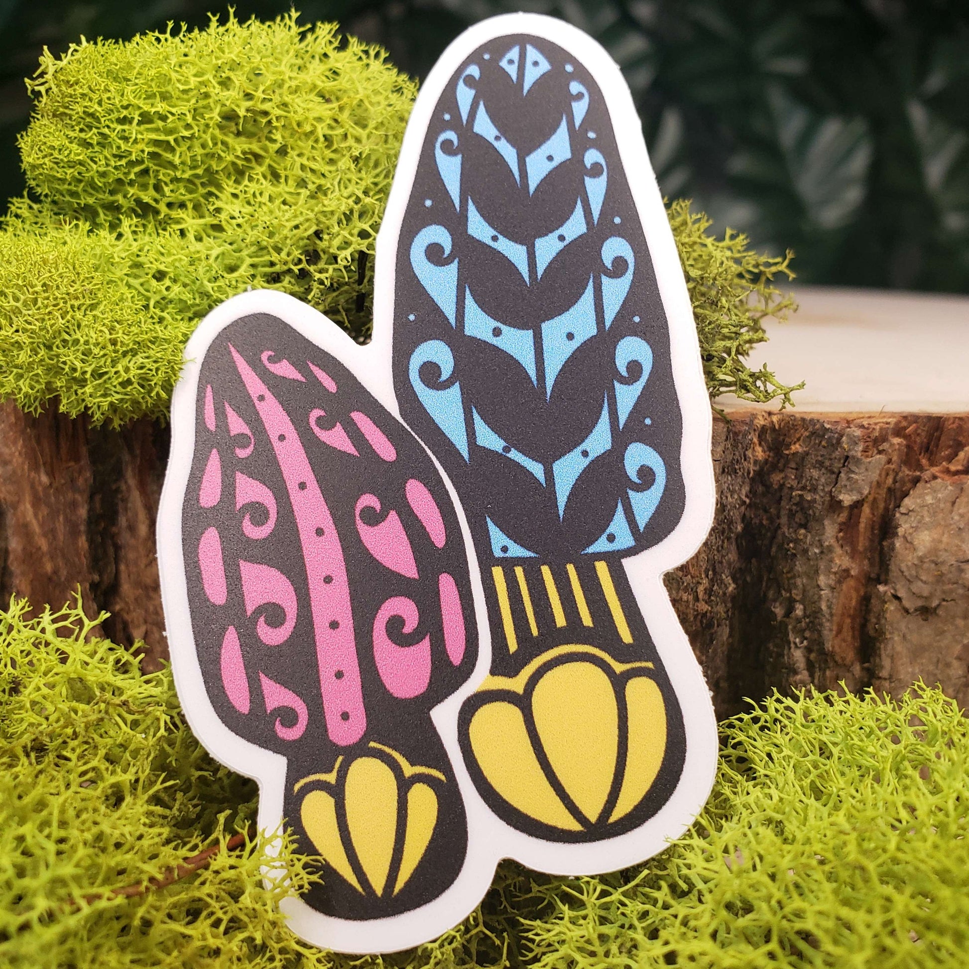 Sticker of pink, blue, and yellow morel mushrooms with stark black line art reminiscent of henna tattoo art. the sticker is sitting on moss and wood with ivy leaves in the background