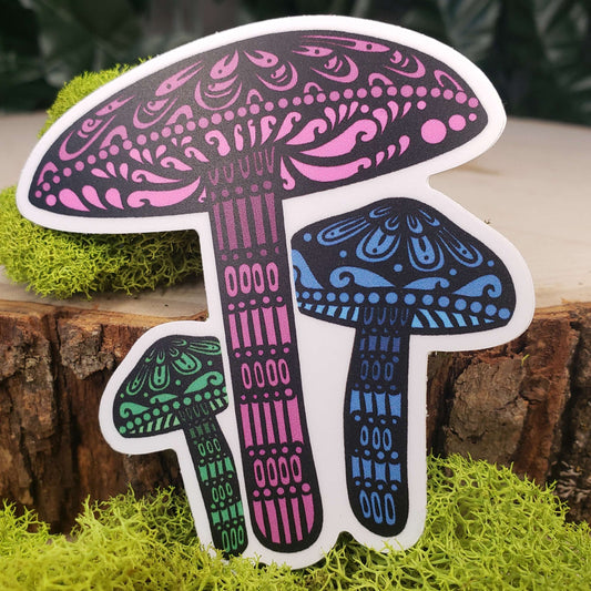 Sticker of green, pink, and blue spring toughshank mushrooms with stark black line art reminiscent of henna tattoo art. the sticker is sitting on moss and wood with ivy leaves in the background
